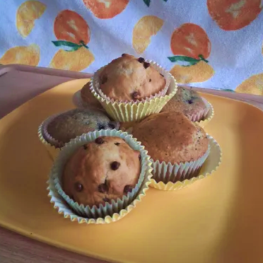 A pile of muffins on a square, yellow plate. There are 3 kinds of muffin, 2 of each in the pile. The background is a lemon themed towel and the table is wooden.